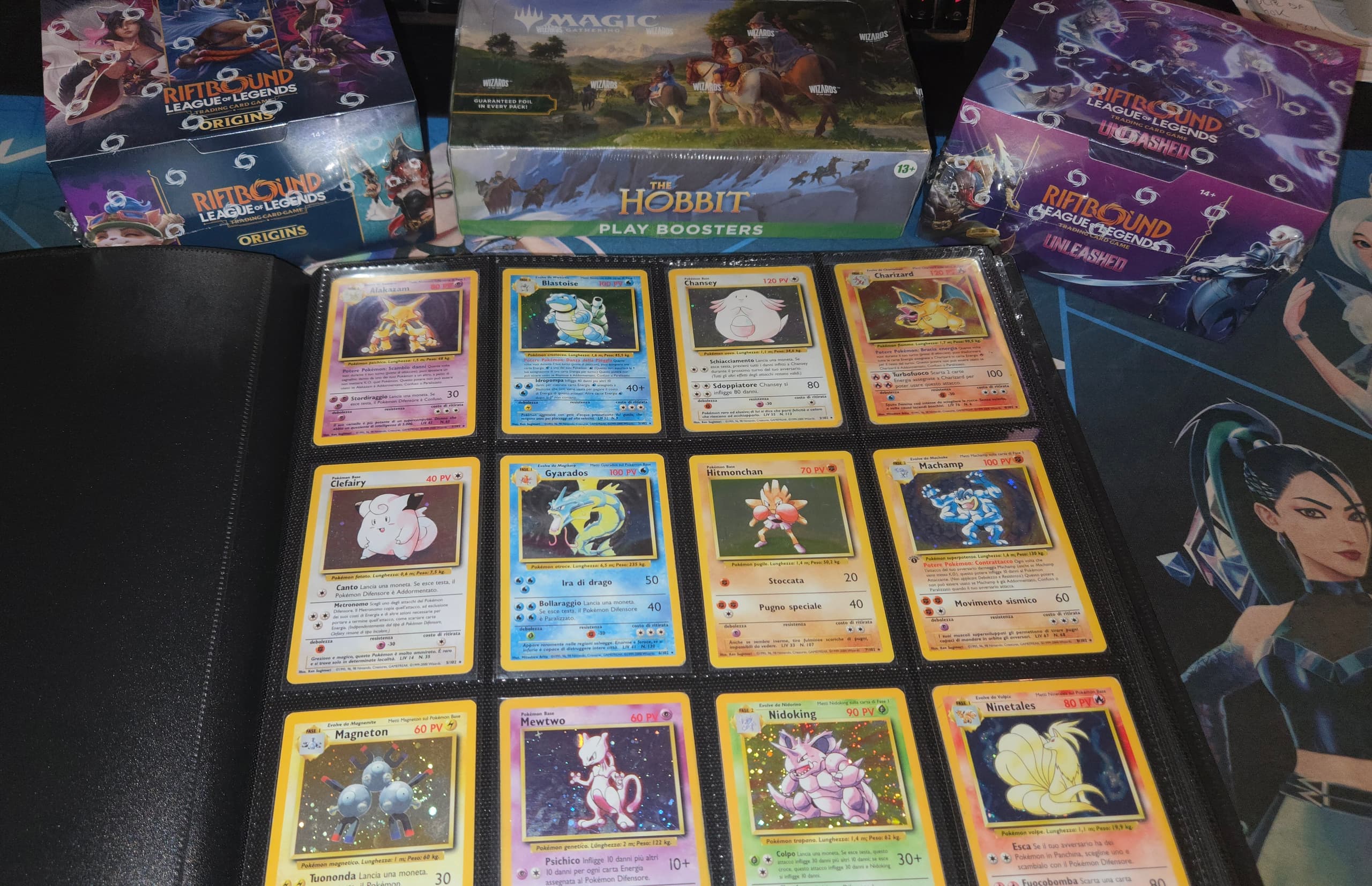 Binder Pokémon holografiche e box sigillati Magic e Riftbound su playmat